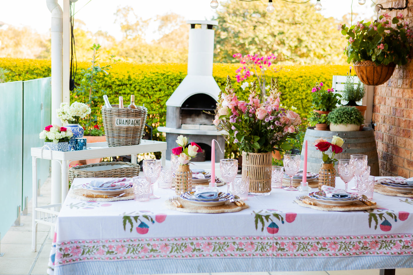 Outdoor table setting with floral tablecloth, plates, and candles on a patio with an oven and plants in the background.