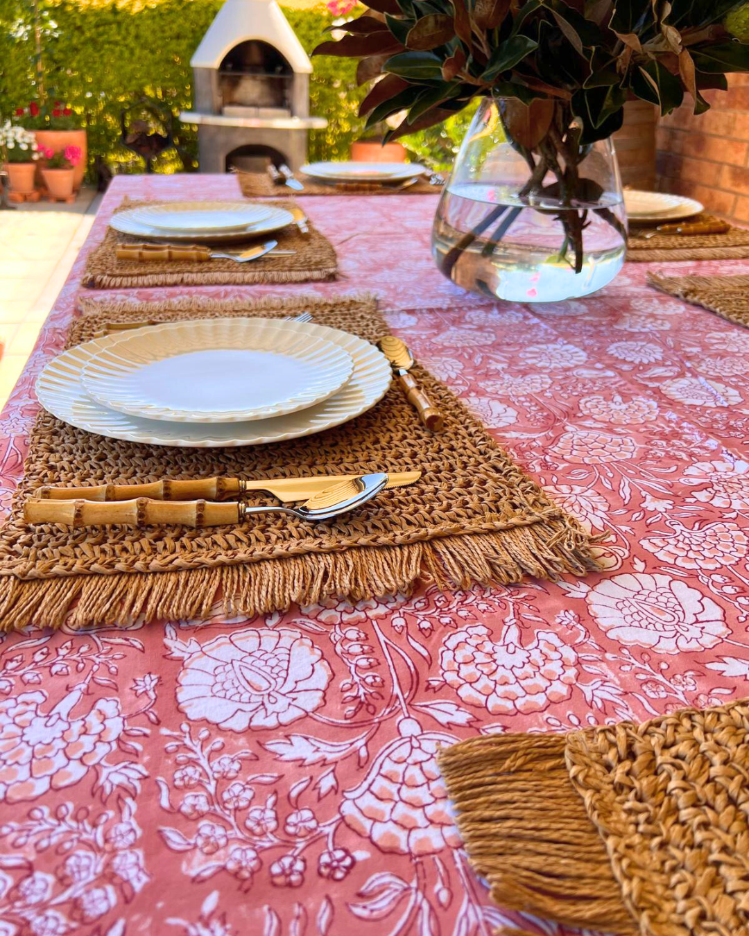 Outdoor table setting with a pink floral Valencia tablecloth, wicker placemats, and decorative elements.