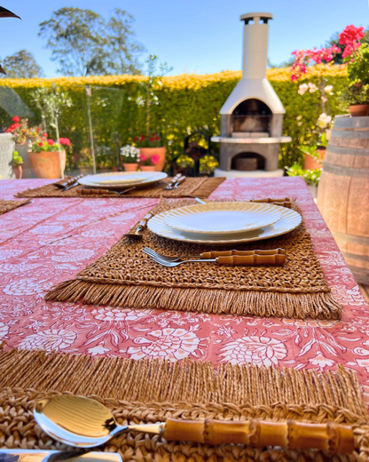 Outdoor table setting with pink tablecloth, brown placemats, and white plates in a garden setting.