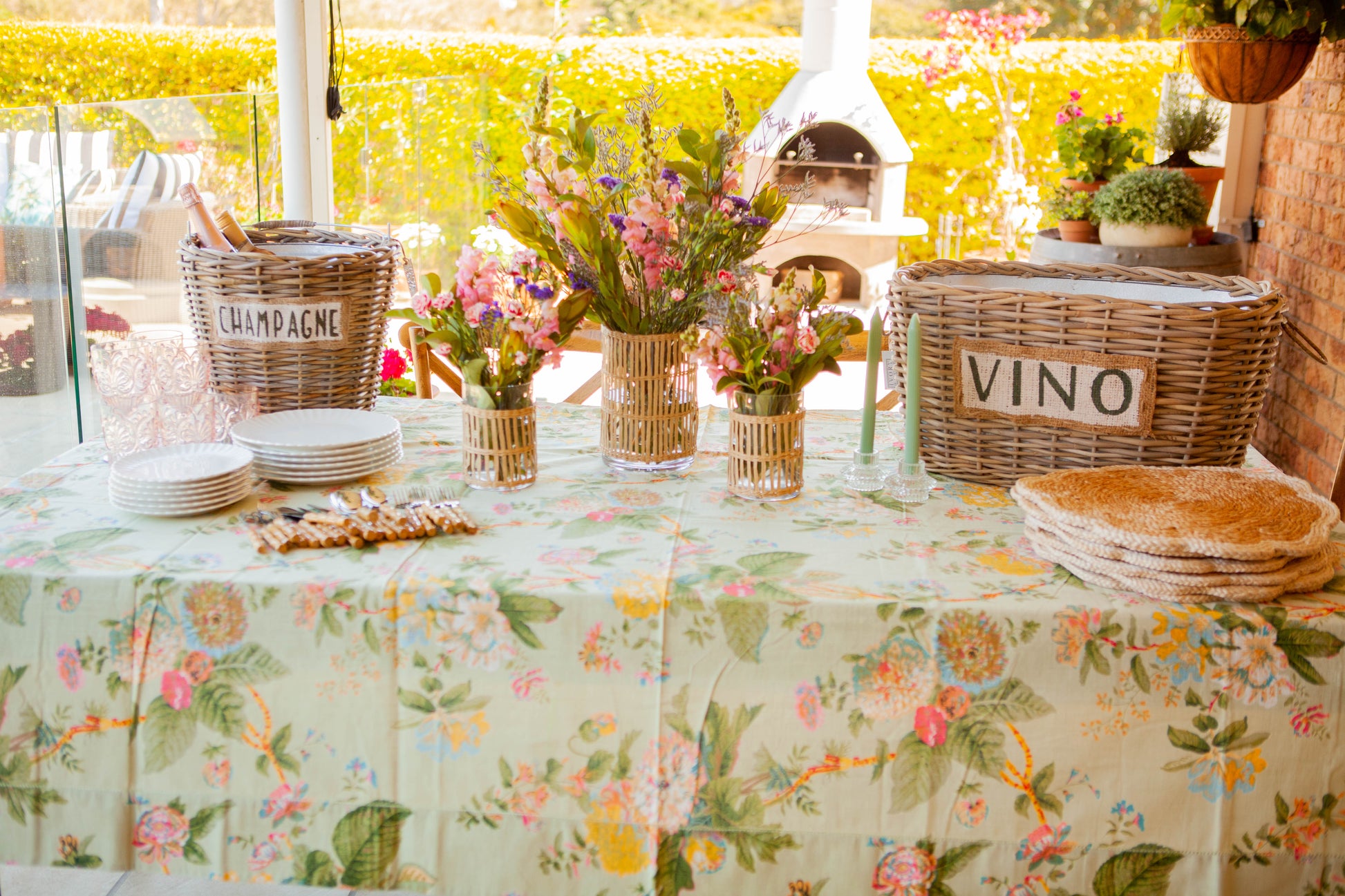 Outdoor table setting with floral tablecloth, wine baskets, and flowers.